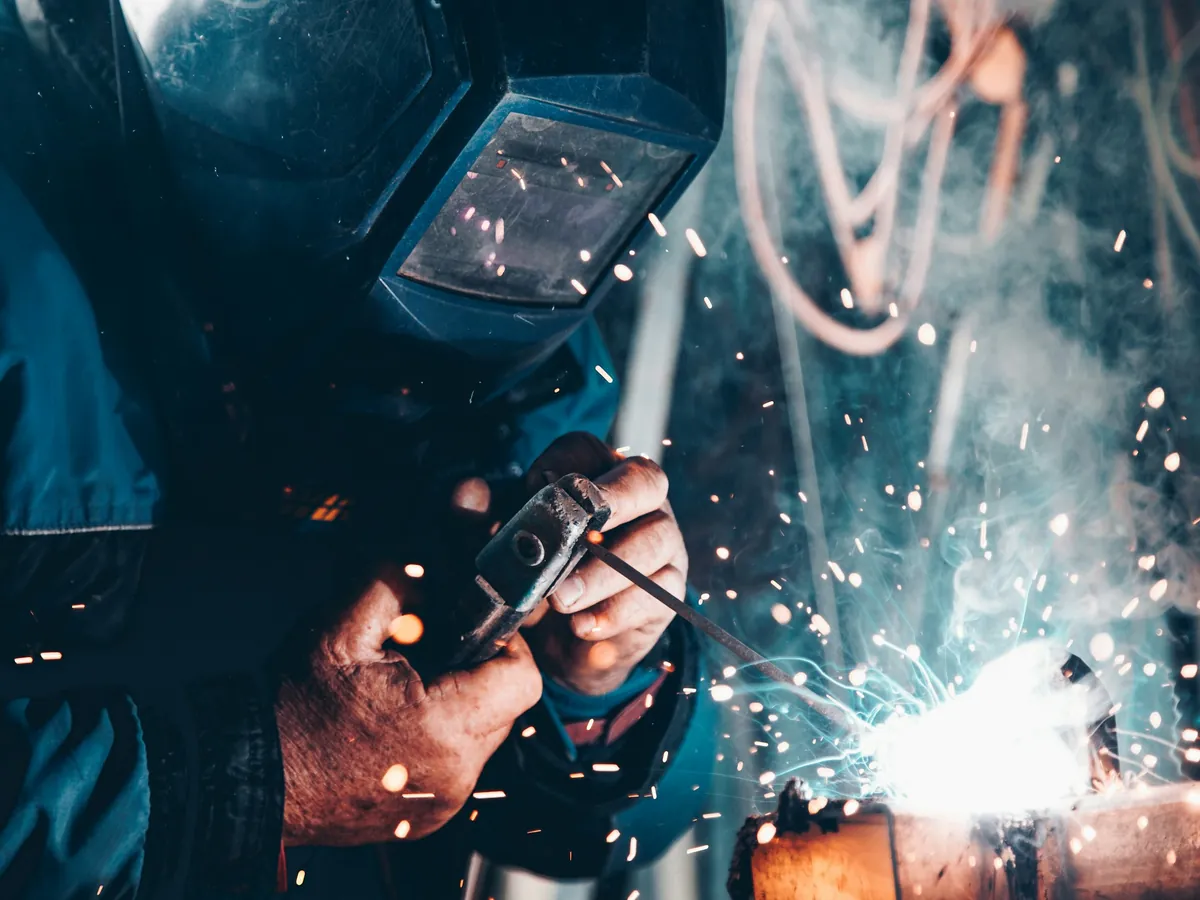 A welder in a helmet stick welding a steel pipe, with sparks flying off the joint